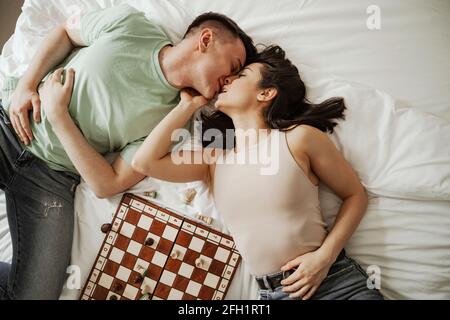 Happy young couple is laying and kissing in bed with chess on it. Young family moving to new apartment Stock Photo