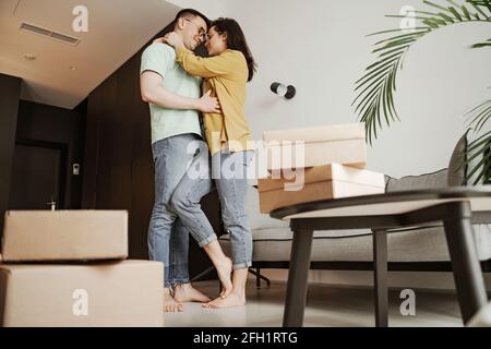 Happy couple is smiling and looking at each other with boxes on flour in new flat. Young family moving to new apartment Stock Photo