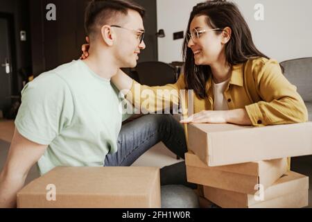 Happy couple is smiling and looking at each other while sitting on flour with boxes at home. Young family moving to new apartment Stock Photo