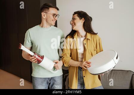 Happy young couple is smiling while looking at each other and holding robot vacuum cleaner at home Stock Photo