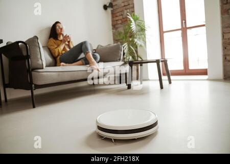 Lovely young woman sitting on couch in living room while robot vacuum cleaner working. Happy family moving to new apartment Stock Photo