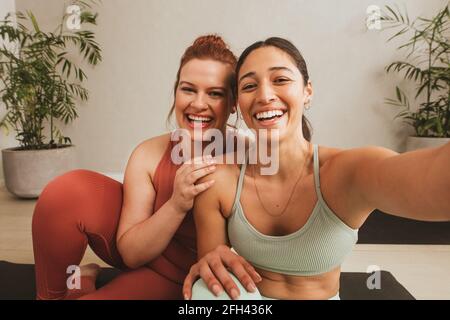 Cheerful female friends taking selife at yoga gym. Women in sports wear taking a break from workout and talking selfie at fitness studio. Stock Photo