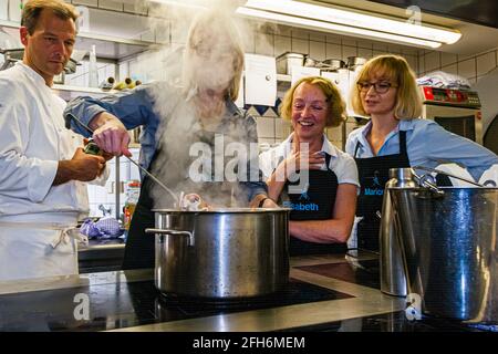 Michelin-starred chef Thomas Kellermann pours an ingredient into a steaming pot with the langostino carcasses – the base for the sauce of his signature dish – in the kitchen of Wernberg Castle, Wernberg-Köblitz, Upper Palatinate, Germany. Journalist Angela Berg (centre left, in the steam) stirs, while participants Elisabeth (centre right) and Marion (right) look on. A peaty, caramel-toned cognac is later added to the sauce, among other ingredients. Stock Photo