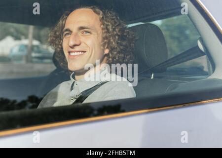 Through window view of smiling young stylish haired male passenger sitting on backseat of modern automobile and fastened with safety belt enjoying tri Stock Photo