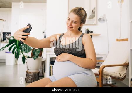Young content expectant female sitting on fitness ball while taking selfie on cellphone at home Stock Photo