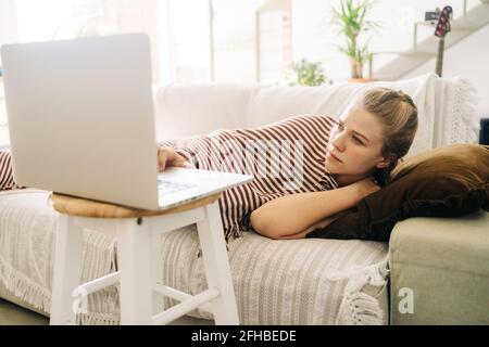 Young exhausted female lying on couch and watching movie on netbook in living room at home Stock Photo