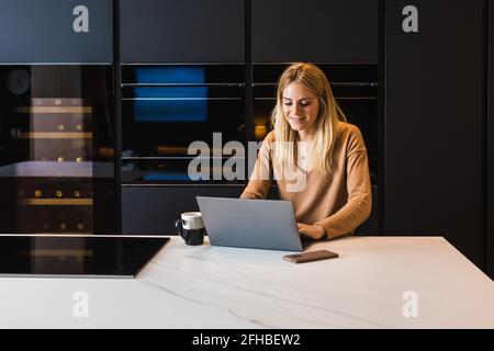 Positive female smiling while talking on video chat via laptop in kitchen at home Stock Photo