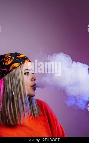 Side view of cool female in street style outfit smoking e cigarette and exhaling smoke through nose on purple background in studio with pink neon illu Stock Photo