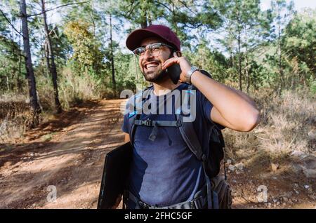 Happy young ethnic male hiker in casual wear and cap with backpack and laptop talking on the mobile phone in lush summer woodland Stock Photo