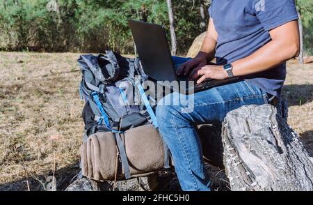 Side view of anonymous nomad sitting on stone near belongings and using netbook while working remotely Stock Photo