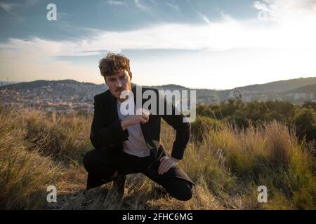 Adult bearded male in coat squatting on faded grass and looking at camera against mounts under cloudy sky in twilight Stock Photo
