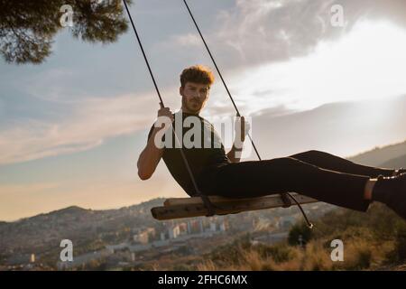 Bearded male in black apparel looking at camera while sitting on swing against mountains in town at sundown Stock Photo