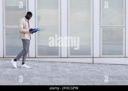 Side view of busy African American male freelancer in smart casual style walking along street and browsing mobile phone Stock Photo