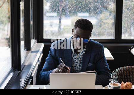 Concentrated African American male entrepreneur writing in notepad while sitting at table with laptop and working in cafe Stock Photo