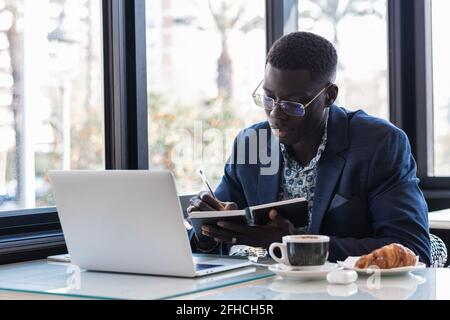 Concentrated African American male entrepreneur writing in notepad while sitting at table with laptop and working in cafe Stock Photo