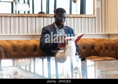 Focused African American male entrepreneur in elegant clothes sitting on sofa in luxury cafe and reading book Stock Photo