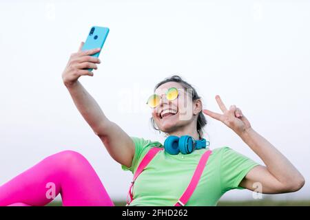 Side view joyful young female wearing bright outfit lying on lush grass taking a self portrait while making the victory symbol in countryside Stock Photo