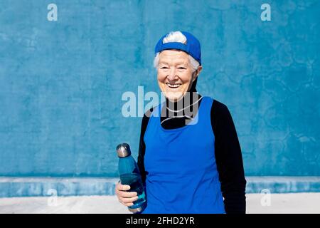 Cheerful mature sportsWoman in activewear and cap standing with water bottle in hand against blue wall in sunny outdoor training center and looking aw Stock Photo