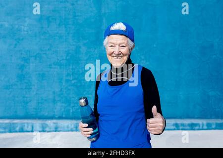 Cheerful mature sportsWoman in activewear and cap standing with water bottle in hand against blue wall in sunny outdoor training center and looking at Stock Photo