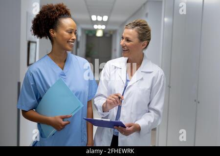 Two diverse female doctors standing in hospital corridor smiling to each other Stock Photo