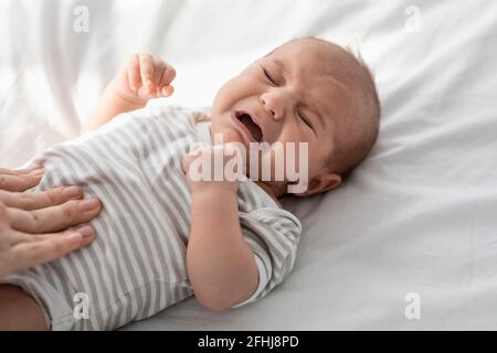 Closeup Portrait Of Crying Little Newborn Baby In Bodysuit Lying On Bed Stock Photo