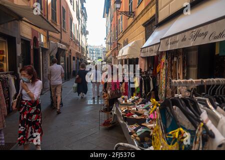 Clothes are for sale outside a store in a narrow shopping street in the pedestrian area of the old town of Rapallo. Stock Photo