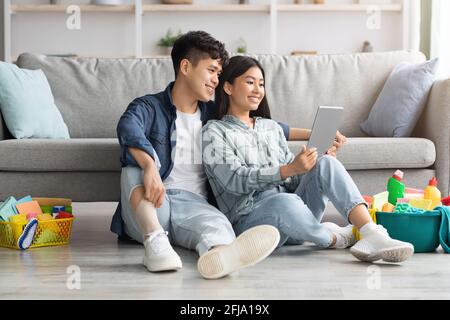 Loving asian couple with cleaning tools resting, using digital tablet Stock Photo