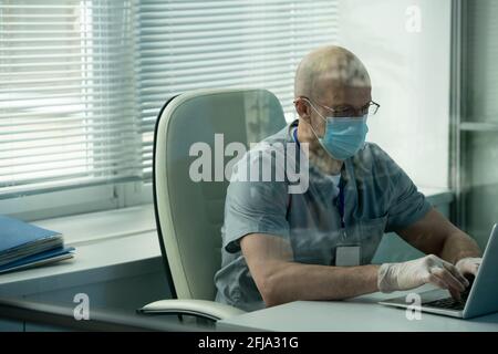 Serious bald surgeon in mask and gloves sitting at desk in office and typing on laptop while working with online records of patient, behind glass view Stock Photo