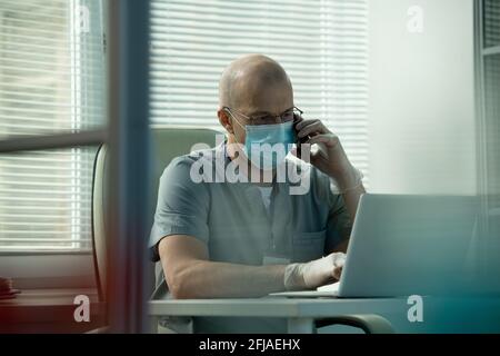 Thoughtful bald medical specialist in scrubs sitting at desk and using laptop while discussing online records with colleague by phone Stock Photo
