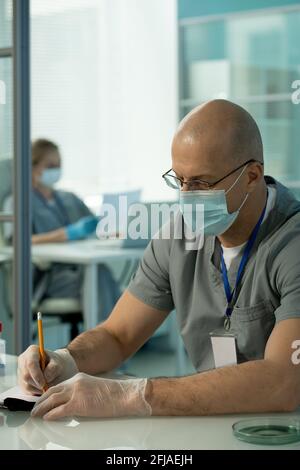 Concentrated middle-aged doctor in mask and gloves wearing badge on neck sitting at table and filling patients record Stock Photo