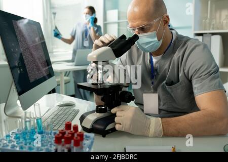 Serious bald man in protective goggles and facial mask focused on medical research sitting at desk with computer and using microscope Stock Photo
