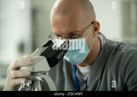 Concentrated lab worker in glasses and mask using microscope while analyzing sample in laboratory Stock Photo