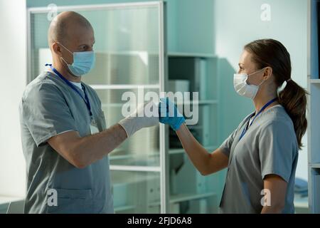 Experienced bald surgeon in scrubs and mask making fist bump with assistant while they preparing for operation Stock Photo