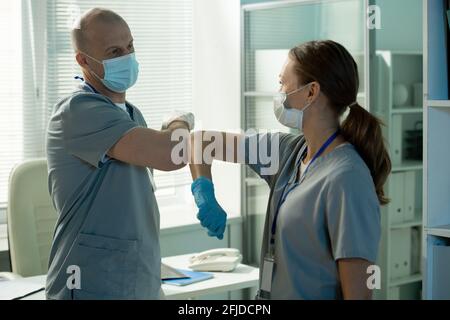 Confident surgeons in scrubs and sterile gloves standing at table in modern office and tapping elbows together while greeting each other Stock Photo