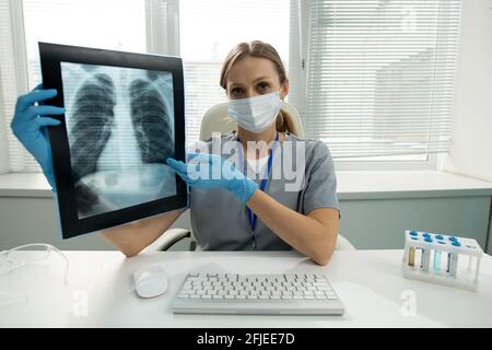 Portrait of female radiologist in mask and gloves sitting at desk with computer keyboard and test tube rack and explaining x-ray image to you Stock Photo