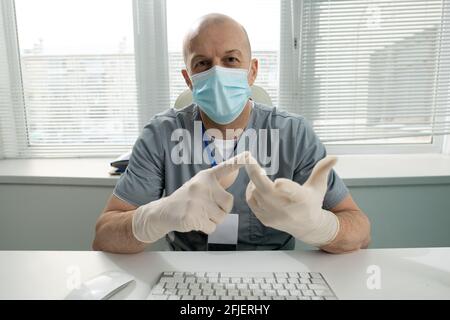 Portrait of bald middle-aged doctor in mask and gloves sitting at table and counting fingers while listing symptoms Stock Photo