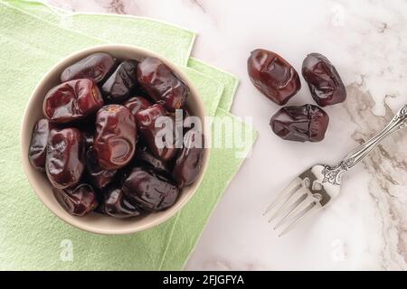 Tasty dried dates in a bowl and fruit fork on a marble surface. Sweet vegetarian, dieting and healthy food ingredient. Organic Iranian dates. Stock Photo