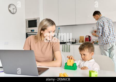 Happy mother working from home using laptop sitting at table with cute kid son. Stock Photo