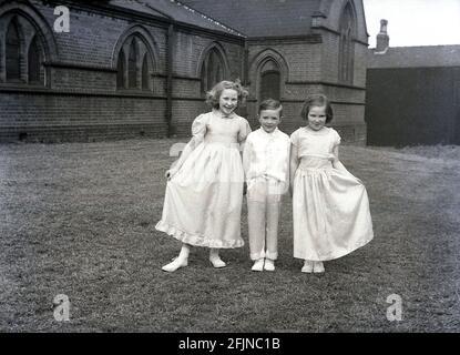 1956, historical, outside in the grounds of church, two young girls and a little boy stand for a photo in their outfits for the traditional May Queen carnival, England, UK. An ancient festival celebrating the arrival of Spring, in Britain in this era, May Day involved the crowning of a May Queen and dancing around a Maypole, activities that have taken place in England for centuries. Selected from the girls of the area, The May Queen would start the procession of floats and dancing. In the industrialised North of England, the Church Sunday Schools often led its organisation. Stock Photo