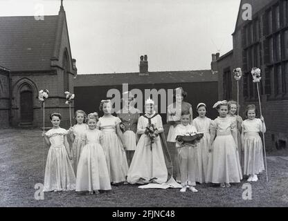 1956, historical, outside in the grounds of church, the town's May Queen standing with her entourage of young girls all in their pretty long gowns, with a solidary little boy holding a cushion, Leeds, England, UK. An ancient festival celebrating the arrival of Spring, May Day involved the crowning of a May Queen and dancing around a Maypole, activities that have taken place in England for centuries. Selected from the girls of the area, The May Queen would start the procession of floats and dancing. In the industrialised North of England, the Church Sunday Schools often led the organisation. Stock Photo