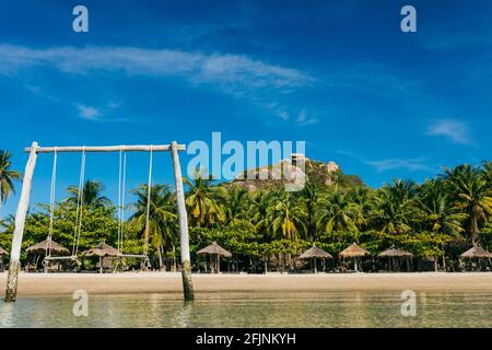 A tropical beach with a wooden swing in the water, a sandy beach on a paradise island, an empty beach with parasols near the coconut trees with a moun Stock Photo