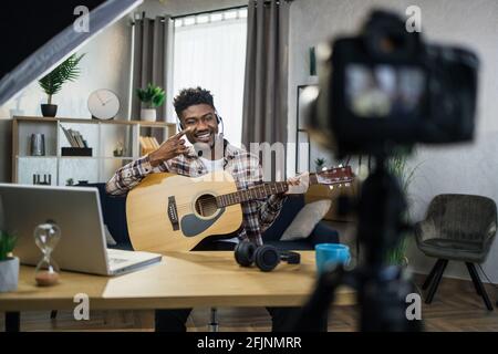 Smiling african blogger playing guitar and filming it on modern video camera. Experienced musician sitting at home and making online tutorial. Stock Photo