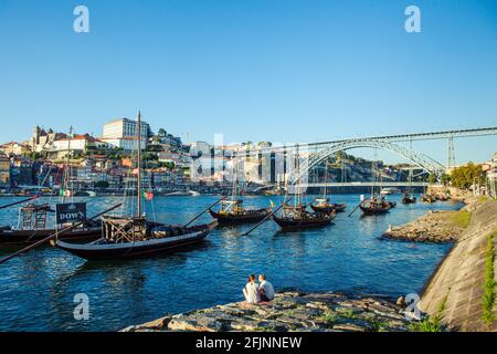 A couple sitting by the river Douro in Porto, with the traditional Port wines  Barges and the Dom Luis bridge in the background, Portugal. Stock Photo