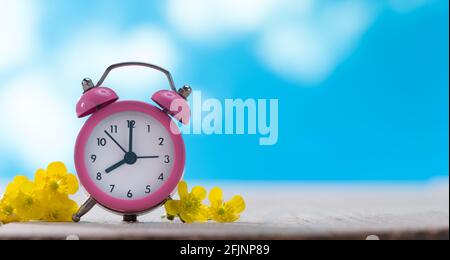 Pink Clock and Yellow Flowers on the Table. Unclear blue sky and white clouds in the background Stock Photo