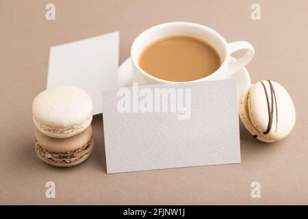 White paper business card mockup with brown and white macaroons and cup of coffee on beige pastel background. Blank, side view, copy space, still life Stock Photo