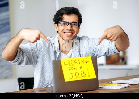 Unemployed positive Hispanic guy with glasses, a student sits at his desk, pointing with his fingers at a sign on a laptop with an inscription looking for a job, looking at the camera, smiles friendly Stock Photo