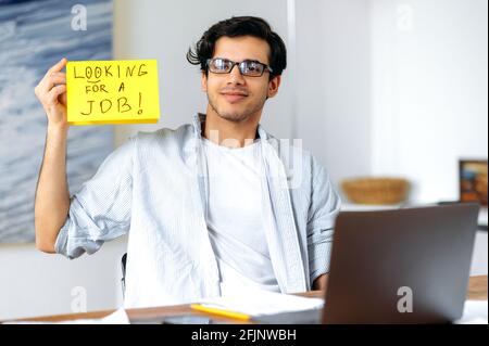 Unemployed young smart hispanic man with glasses, student or freelancer, sits at table, holds sign with inscription looking for job, hopes for help, asks for support, looks at camera friendly Stock Photo