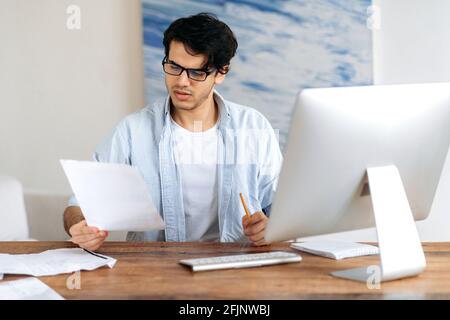 Concentrated confident successful handsome hispanic man with glasses, freelancer, student or businessman working or studying rat the desk with computer, looking through business documents Stock Photo