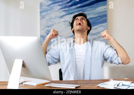 Joyful successful young Hispanic man, businessman, manager or freelancer, in stylish clothes, working at the computer, happy with victory, win or big profit, gesturing with his hands, shouting happily Stock Photo