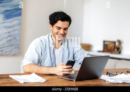Happy young smart hispanic guy, freelancer, manager or student, working or studying at laptop, wearing casual clothes, using a smartphone, texting with a colleague, client or classmate, smiling Stock Photo
